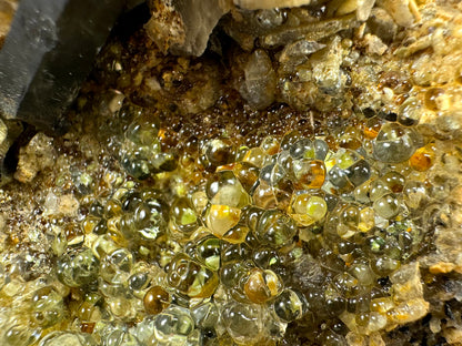 Detail of botryoidal hyalite with good transparency and a slightly green-yellow tint. The matrix visible beneath is multi-colored tones of yellow, orange, brown, and black.