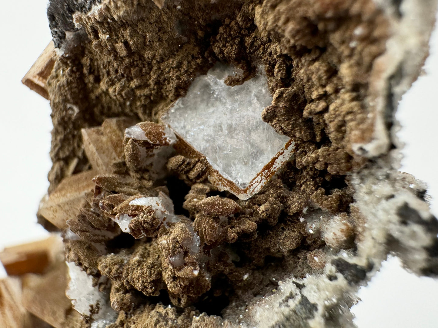 Detail of a translucent white hydroxyapophyllite crystal with an almost cross-hatch pattern to the surface, and brown spots of lizardite along the side faces. Below to the left is a cluster of stacked scalenohedral calcite crystals, mostly covered in the brown mineral.