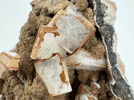 Detail of several translucent white hydroxyapophyllite crystals, with three of the square tabular crystals intersecting. They have a light cubic of crosshatch pattern visible in the inclusions or on the surface. There are brown spots of lizardite along the side faces, but the main square faces are. Below to the left is a cluster of stacked scalenohedral calcite crystals, mostly covered in the brown mineral.