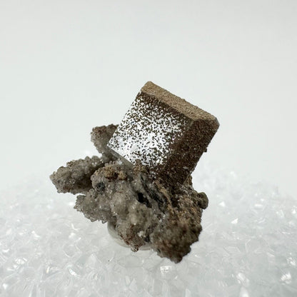 Close view of a square tabular crystal of hydroxyapophyllite, on bunchy off-white druzy. The crystal tilted so light reflects off the main square face, highlighting the small round spots speckling its surface. They coat the top and right edges abut are more dispersed on the main face.