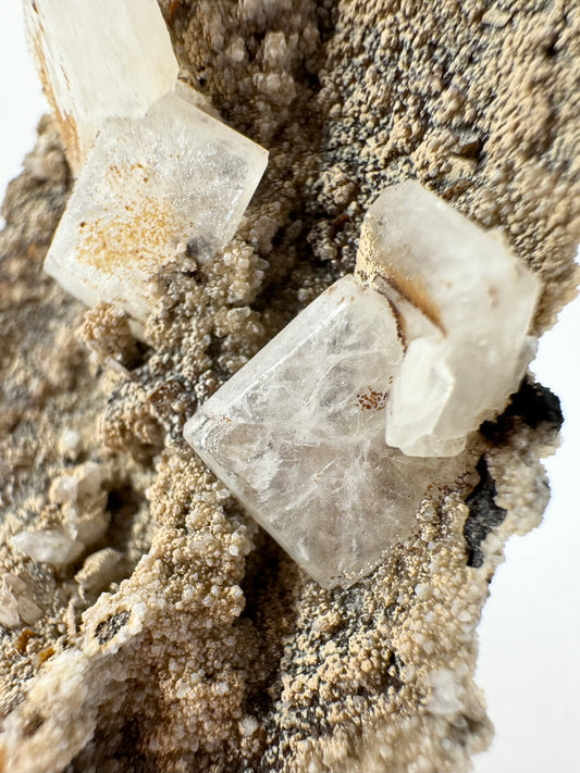 Detail of a translucent white hydroxyapophyllite crystal with an almost spray-like pattern of wispy inclusions out from the center. The surrounding matrix has a sandy texture coated in light lizardite