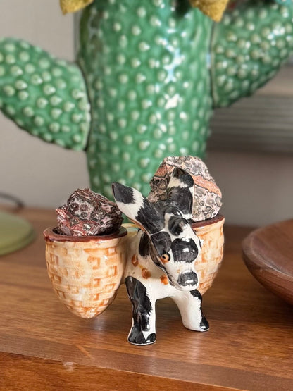 A retro ceramic mule with baskets on each side, each filled with a piece of leopardskin jasper rough. There is a ceramic cactus in the background.