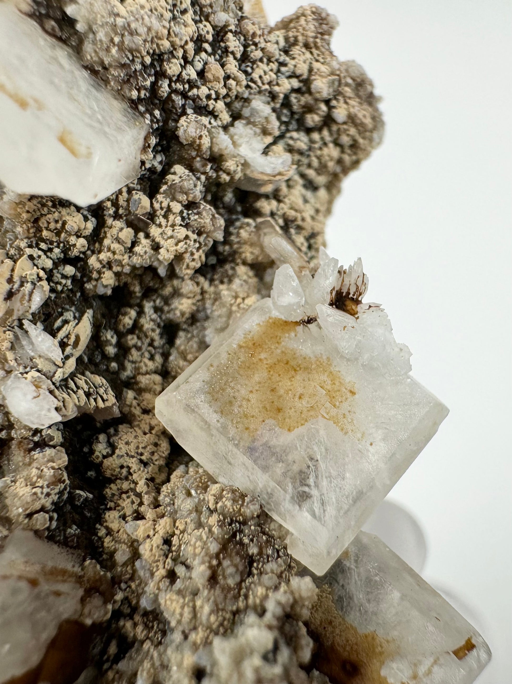 Detail of a translucent white hydroxyapophyllite crystal with a cluster of scalenohedral calcite crystal on its corner. The matrix is textured like cauliflower, dark with light tan lizardite covering the tops of bumps.