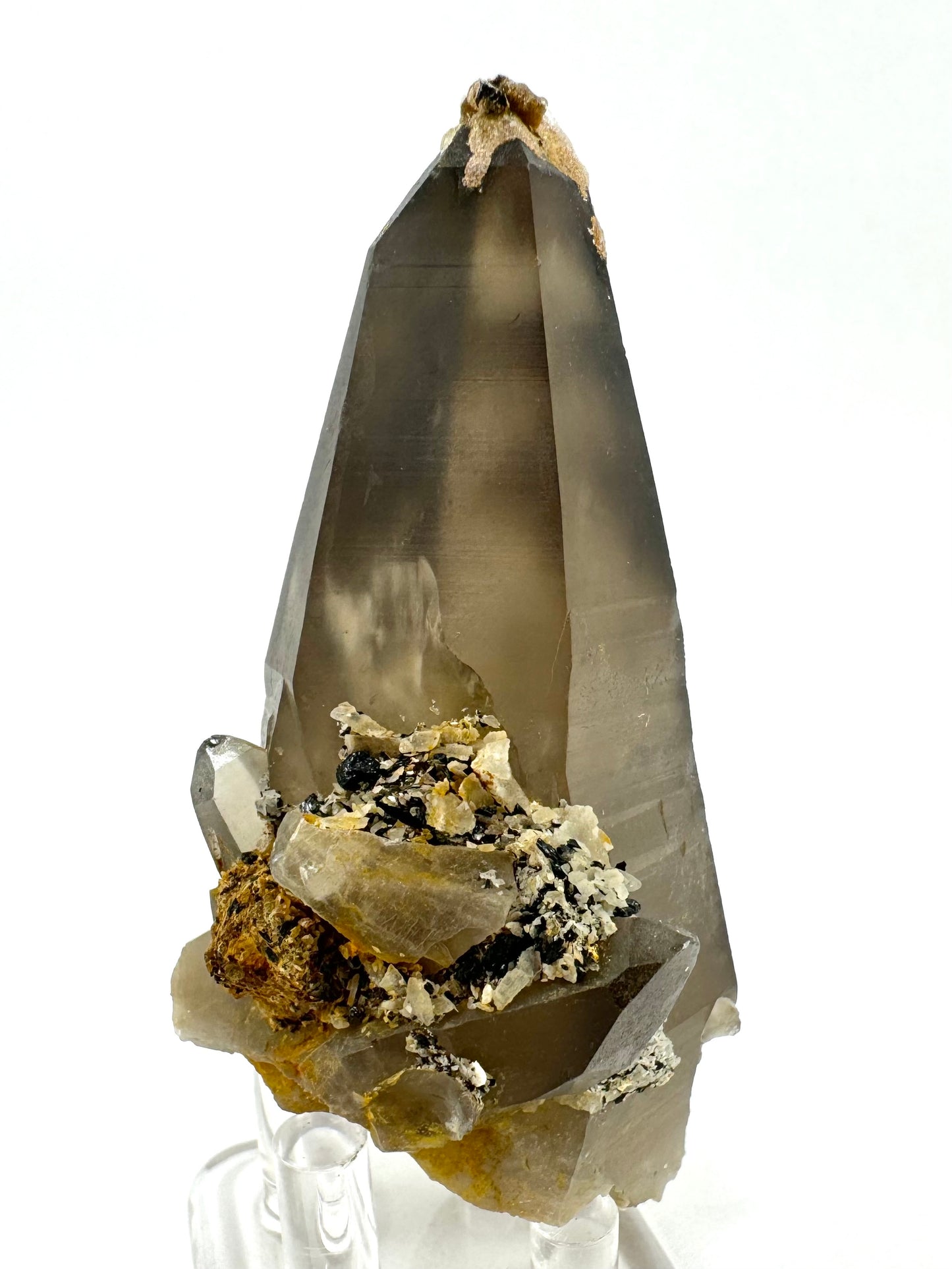 Another angle showing light through the quartz. The body color is fairly light brown and translucent through this side, with a feather inside visible catching light. There is a cluster of smoky quartz and irregular black and white crystals on the base.