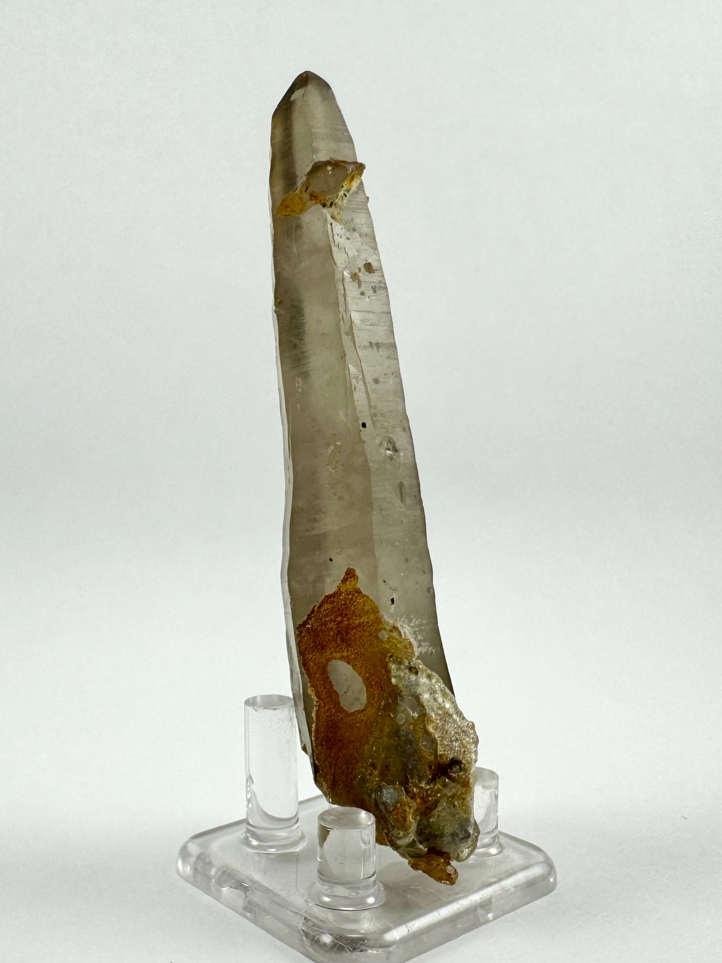 Full view of a spindly long thin quartz prism, with striated faces and a lighter smoky quartz coloration. There is an irregular crystal attached near the tip, and a cluster of minerals on the base. There is an orange-brown tinted overgrowth on the base as well.