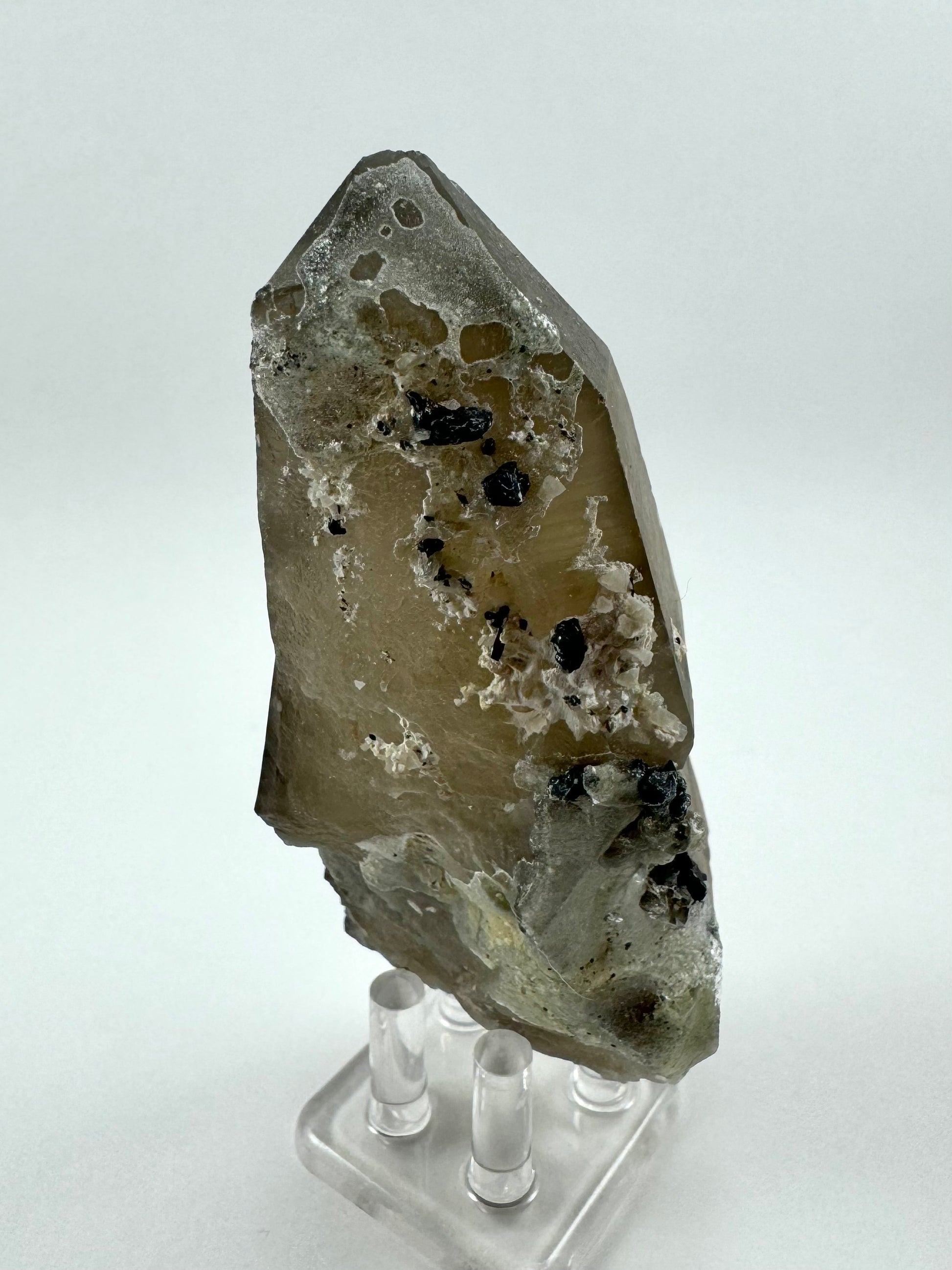 The first of three lighting images: a thick blocky smoky quartz point with white hyalite crust on one side, coating the top and bottom in a cheese-hole pattern. There is some web-like white mineral growth on the center of the quartz. Irregular black schorl crystals are sprinkled throughout.