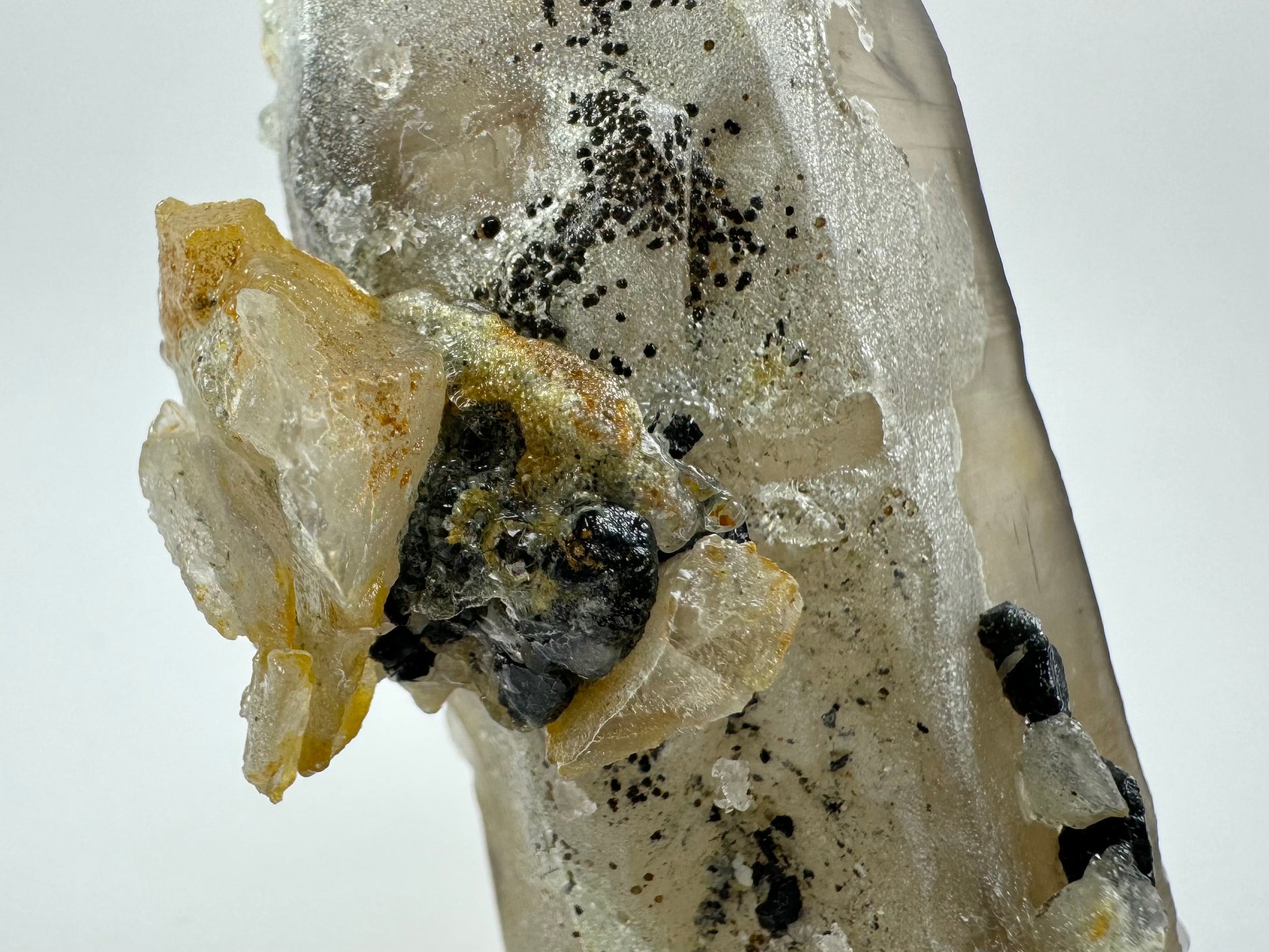 Detail view of the mineral cluster on the smoky quartz, composed of shards of smoky quartz and black schorl fused together and coated in hyalite. There is a cluster of small black shiny spherical minerals spotting the surface of the hyalite. Some of these spheres are broken and appear hollow.