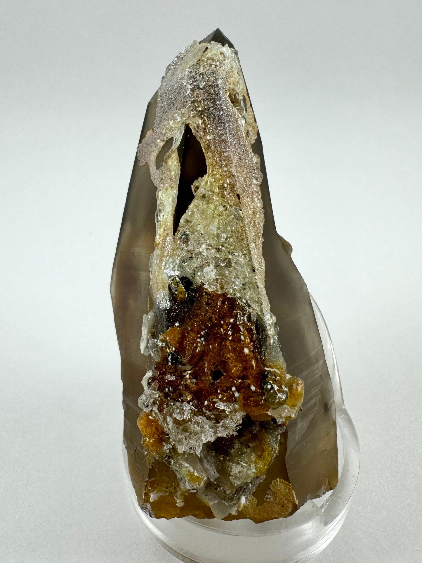 View of the point looking at the side coated in hyalite and with an irregular cluster of minerals near the base. There is black schorl and orange-brown iron staining under glossy clear hyalite.