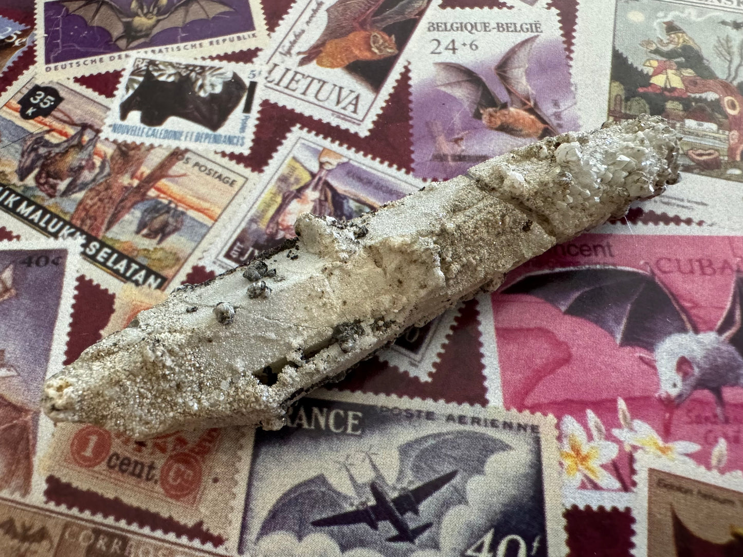 A closer view of the quartz point, with a sandy white crust and irregular white crystals all over it. There are also small black minerals sprinkled on the surface.