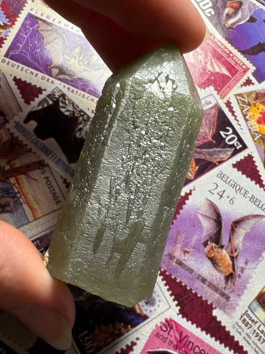 Full view of a stubby hedenbergite quartz point, well-formed straight point with base cleanly broken. It has a light greenish-gray color. It is tilted to reflect light off its surface, highlighting complex sprouting growth forms and small lines etching the surface.