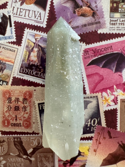 Full view of a hedenbergite quartz point, well-formed slightly tapered point with tip intact and base cleanly broken. Light greenish-gray color deepens closer to the tip. It has small white crystals sprinkled across the surface.