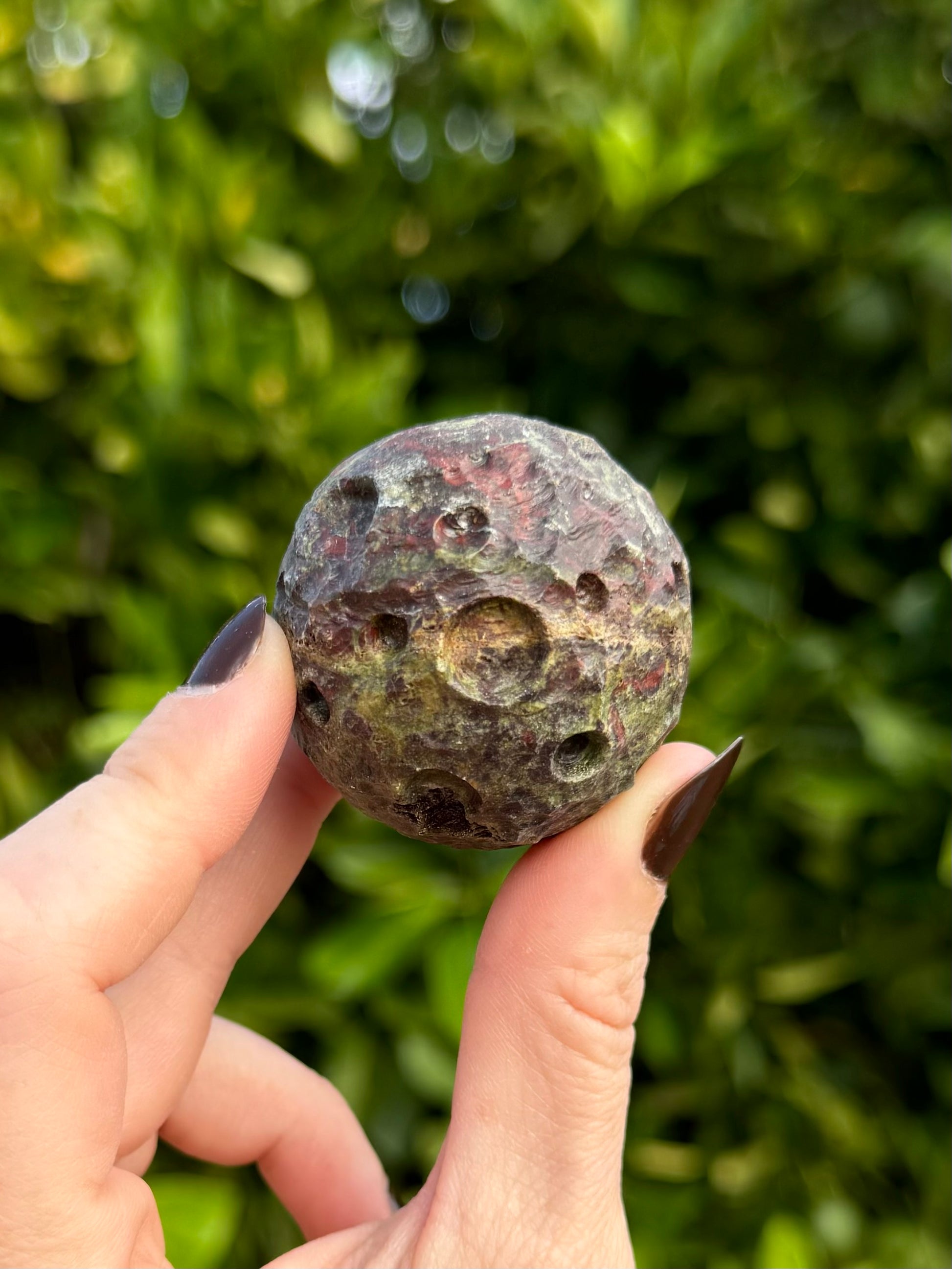 A jasper moon held between thumb and finger to show scale, as well as its colorful combination of green and red with a light streak through the center