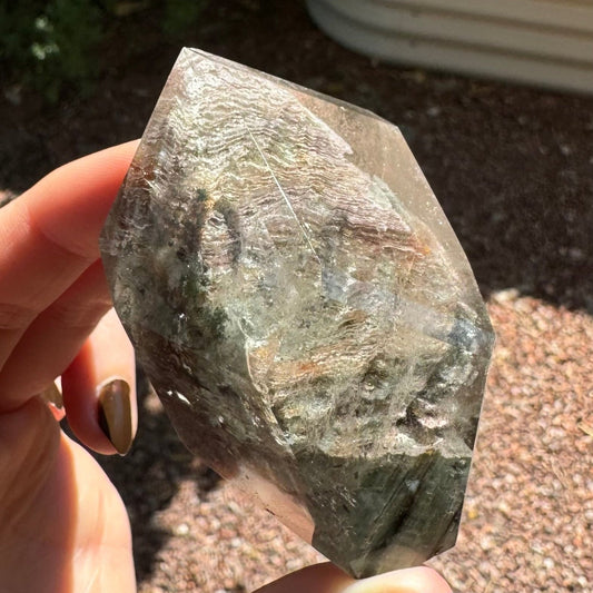 A polished piece of garden quartz held between finger and thumb for scale, in sunlight to highlight internal features. The freeform is blocky, with a flat base and coming to a point at the top back of the piece. It is filled with pale thin layers of inclusions.