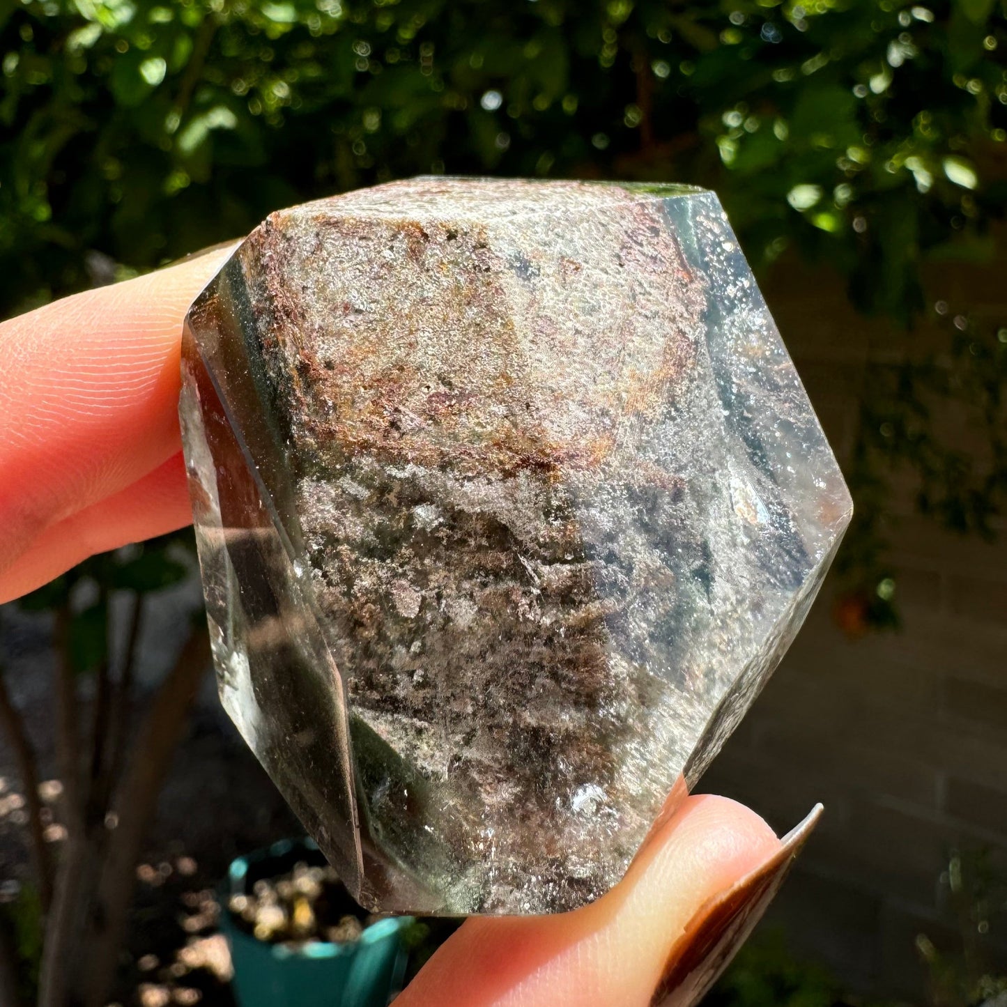 A polished piece of garden quartz held between finger and thumb for scale, in sunlight. The freeform is blocky, cut so three faces come to a point in the center of a large opaque inclusion. The inclusion goes through the piece, and appears reflective and light inside the quartz.