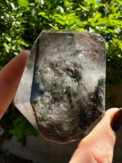 A polished piece of garden quartz held between finger and thumb for scale, in sunlight to highlight internal features. There is a thin dark green layer of chlorite through the piece, wispy colorless inclusions through a lot of it. At the bottom are dark moss-like inclusions reaching up