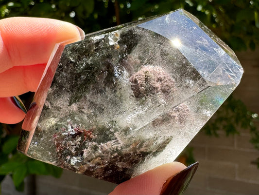 A freeform polished piece of garden quartz with a purple-pinkish tinted large spherical chlorite inclusion, nestled in dark green inclusions with wispy colorless inclusions around it.