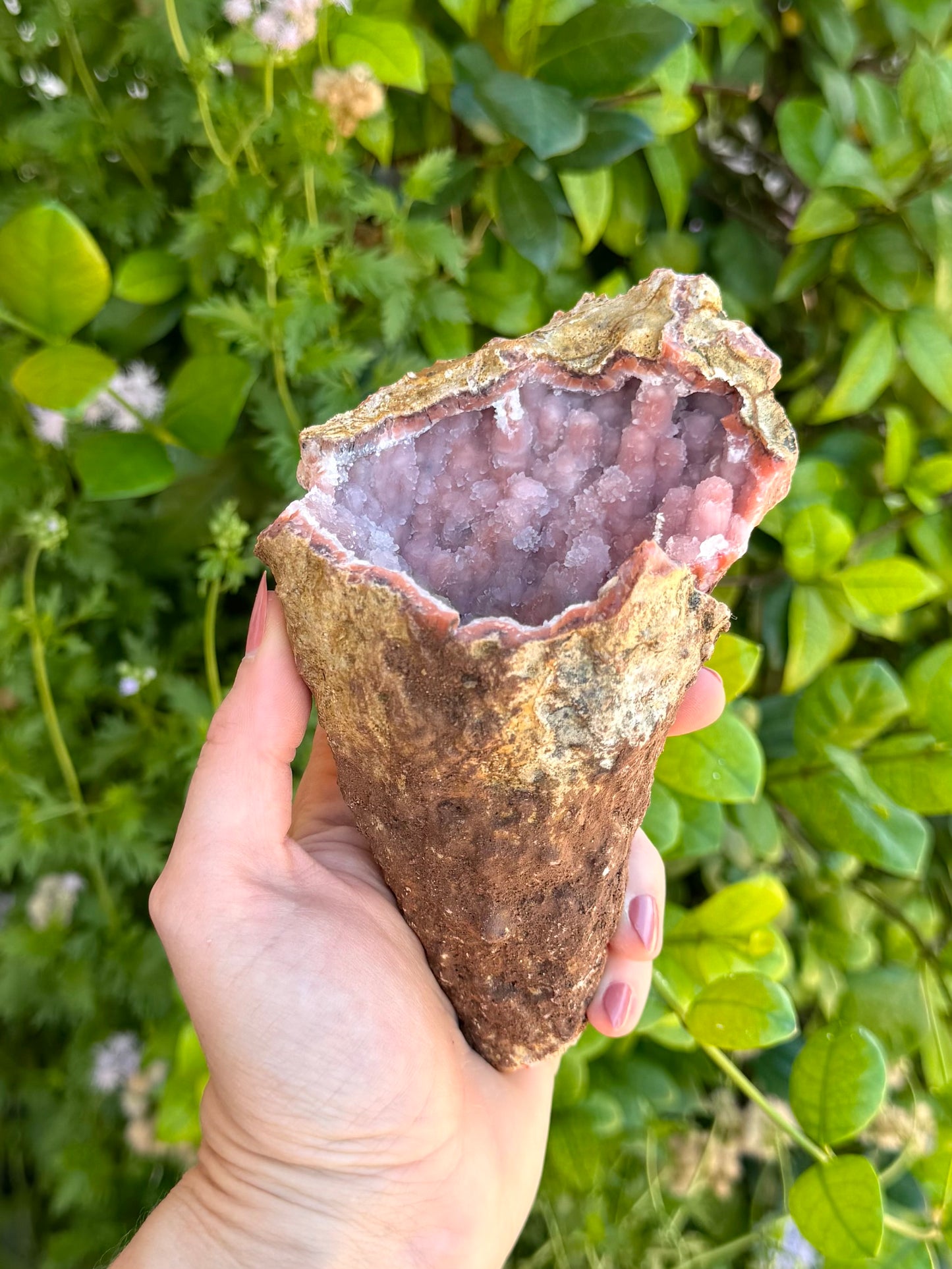 A large conical geode held in the hand like an ice cream cone, in indirect light. The crust of it is a thin rough brown rock, with a darker brown dirt-like covering on the bottom half hiding a repair.