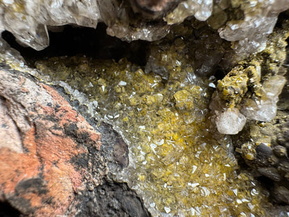 Detail image showing a pocket in the folds of the surface of the specimen, with botryoidal iron oxides covered with thick yellow wulfenite, and a layer of colorless calcite crystals. The crystals are broken in cross-section along the red rock on the lower left, appearing a black bumps with a rim of yellow and white crystal.
