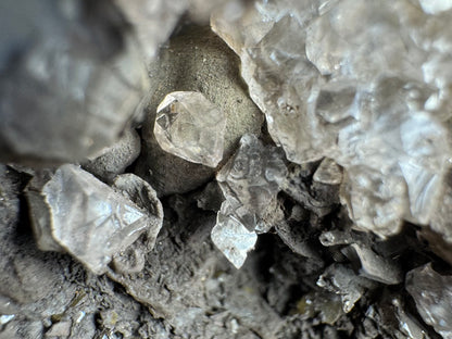 Very close detail image of a sphere of coronadite with a colorless calcite crystal on the surface, surrounded by calcite above and with a craggy black surface below it.