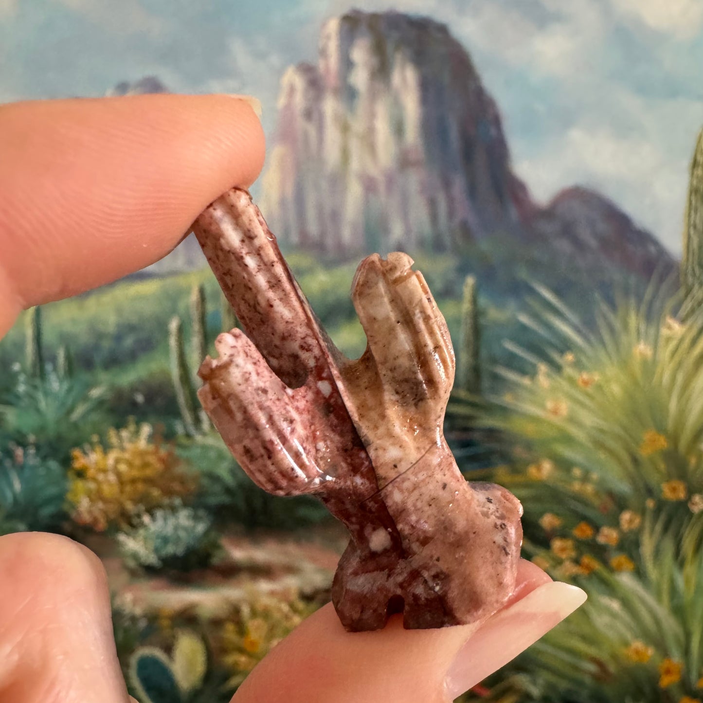 A very small carved saguaro cactus held between finger and thumb. The saguaro has two arms, and the end of each and the main stem are all topped with a flower. It has a bold dark red line through it.