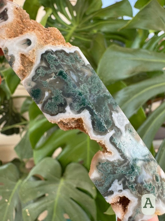 Option A, detail of a moss agate tower with large cavities. The material has mossy green formations lined with translucent chalcedony, then white. It is rough and light brown on the unpolished outside.