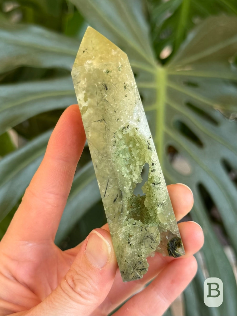 Option B, a four-sided towe carved from translucent green prehnite with small botryoidal growth visible in a cavity through the tower and part of its base.