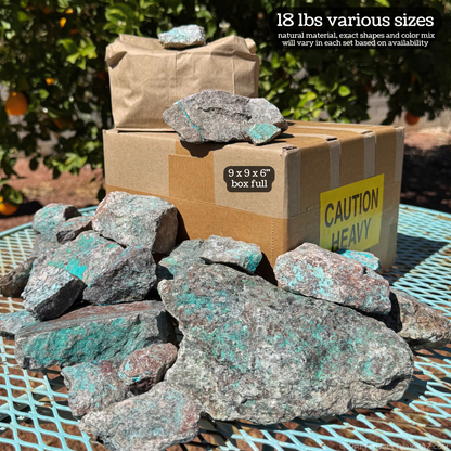 A cardboard box with a pile of various sizes of rough rock spread on and around it on a turquoise-colored table. The image is labeled 18 lbs various sizes, natural material, exact shapes and color mix will vary in each set based on availability. The cardboard box is labeled 9 by 9 by 6 inch box full, and has a "caution: heavy" sticker.